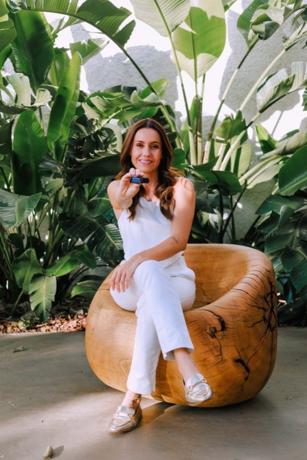 Larissa Buainain segurando um frasco de óleo essencial, sentada em um banco de madeira entre plantas tropicais.